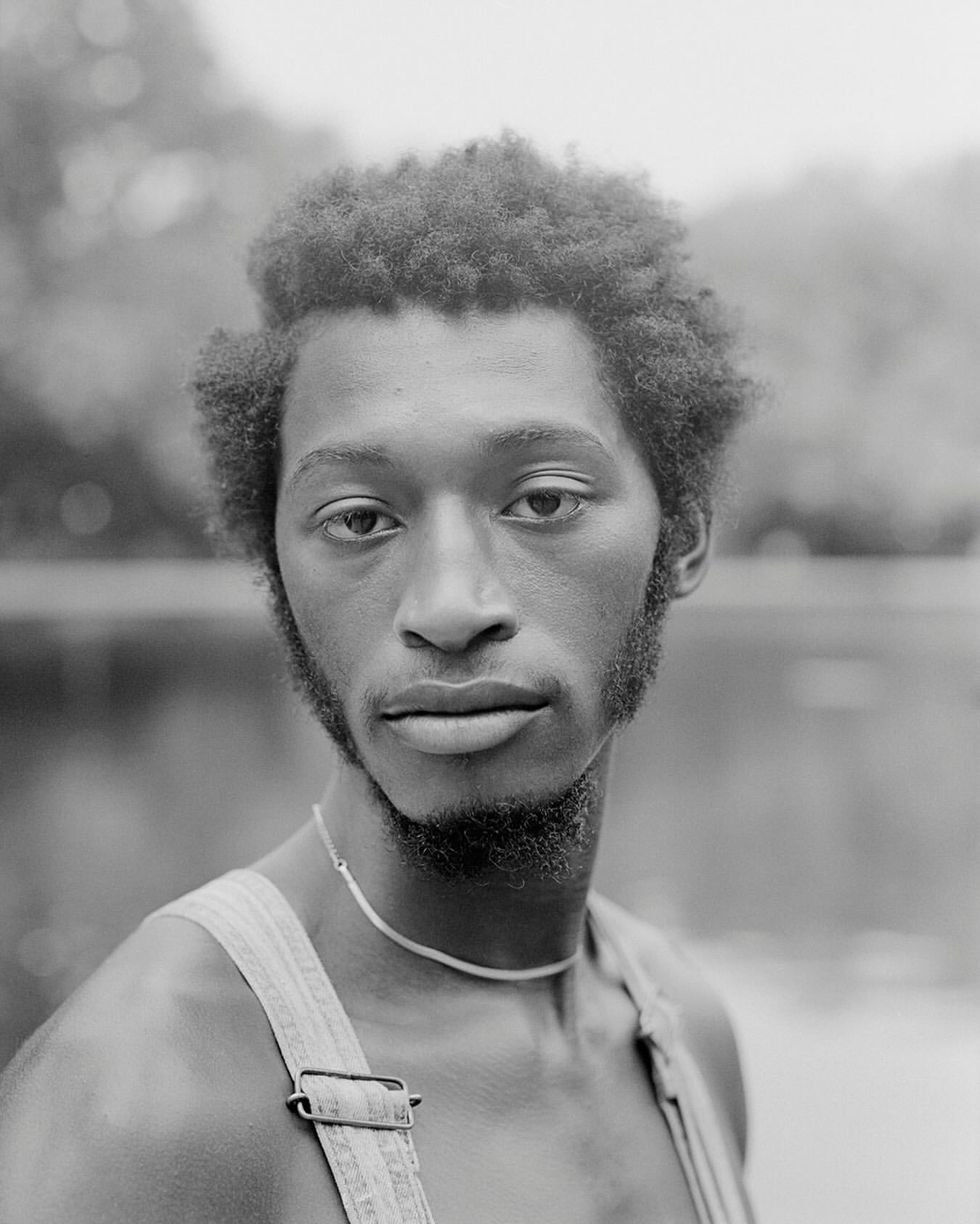 Black and white portrait of a young adult Black man with curly hair, wearing overalls, and standing near a body of water and trees.