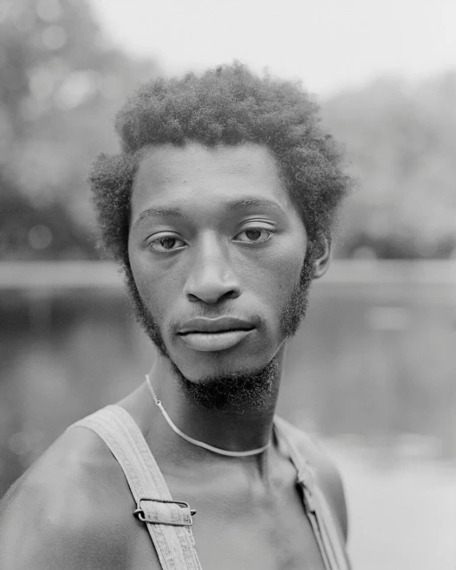 Black and white portrait of a young adult Black man with curly hair, wearing overalls, and standing near a body of water and trees.