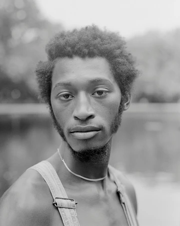 Black and white portrait of a young adult Black man with curly hair, wearing overalls, and standing near a body of water and trees.