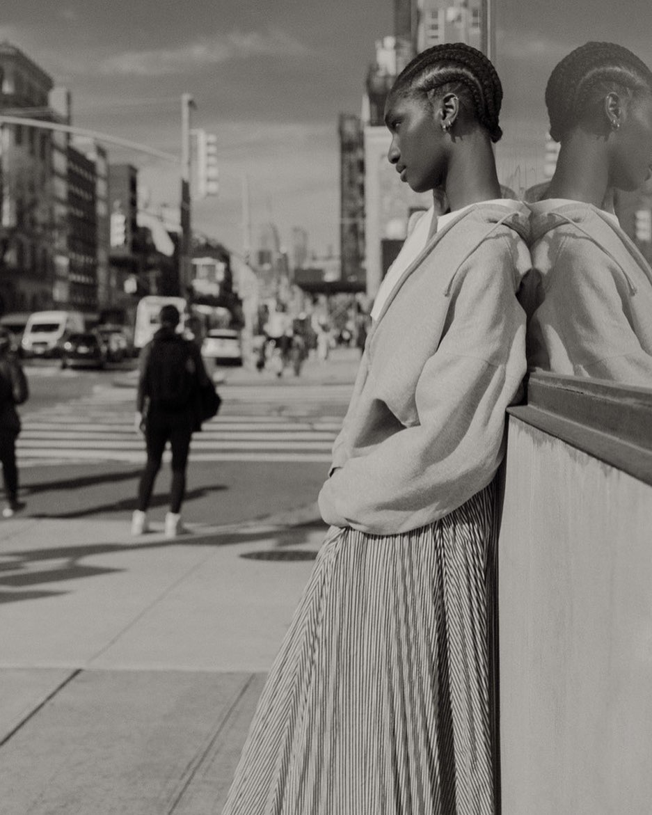 Black and white street style shot featuring an African-American woman in a pleated skirt leaning against a reflective surface in the city.