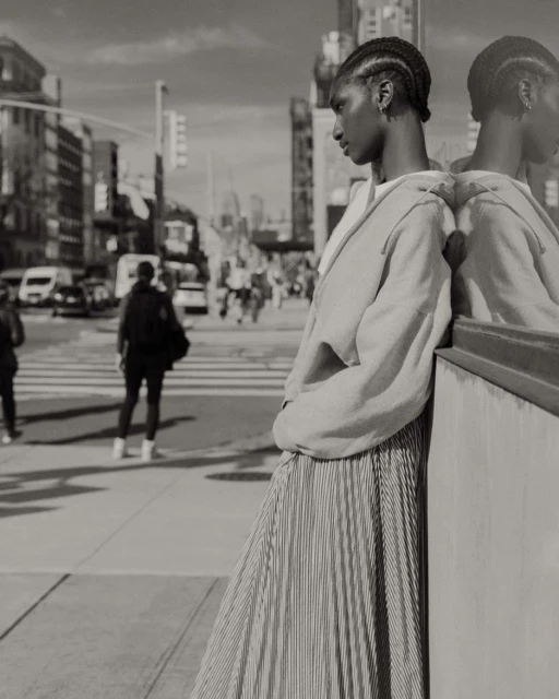 Black and white street style shot featuring an African-American woman in a pleated skirt leaning against a reflective surface in the city.