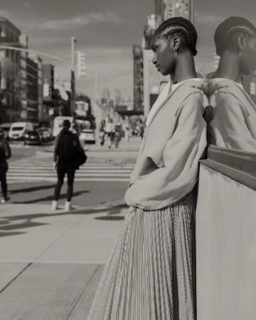 Black and white street style shot featuring an African-American woman in a pleated skirt leaning against a reflective surface in the city.