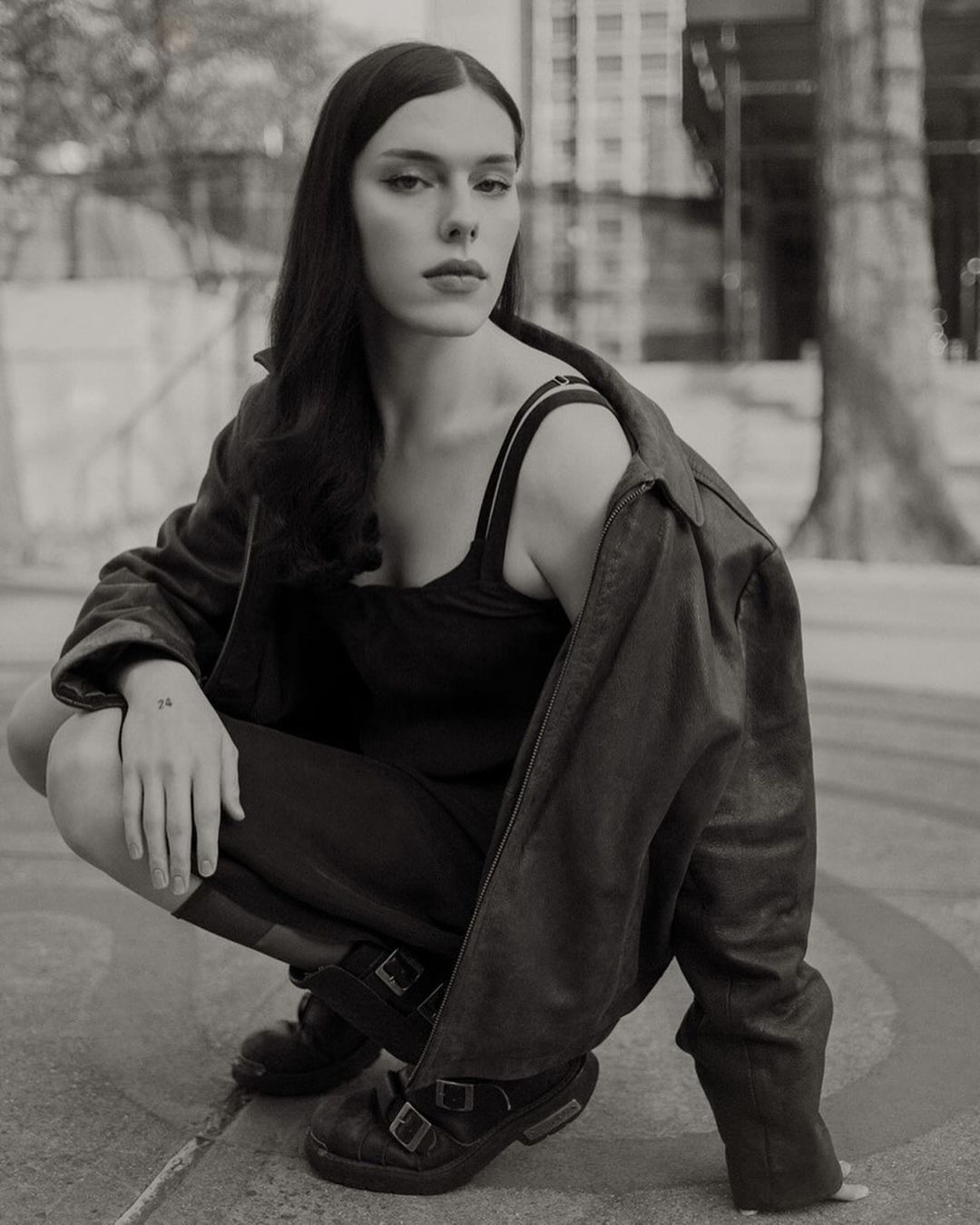 Black and white portrait of a woman in a jacket crouching in an urban setting with buildings in the background, captured at eye level.