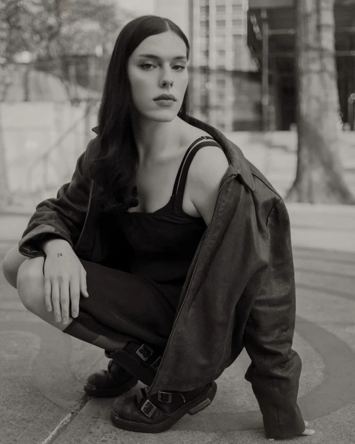 Black and white portrait of a woman in a jacket crouching in an urban setting with buildings in the background, captured at eye level.