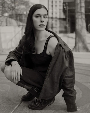 Black and white portrait of a woman in a jacket crouching in an urban setting with buildings in the background, captured at eye level.