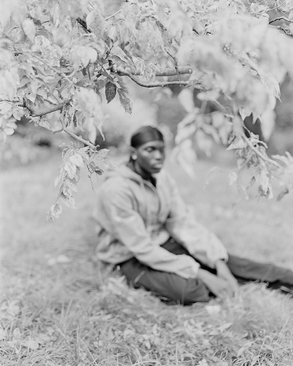 Black and white photo of an African American man sitting beneath tree branches, wearing a jacket and durag in an outdoor setting.