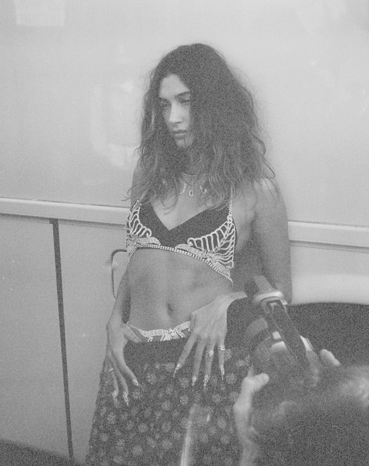 Monochrome portrait of a woman with long hair, wearing a decorative bra and skirt, posing indoors in front of a white wall.