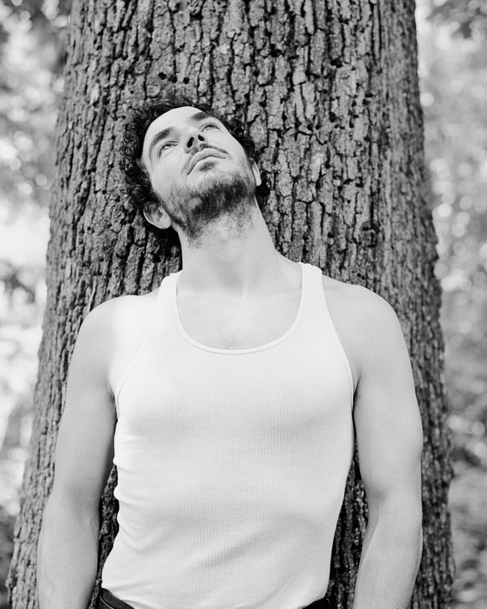 Black and white portrait of a man leaning against a tree trunk, looking up with a slightly tilted head in an outdoor setting.