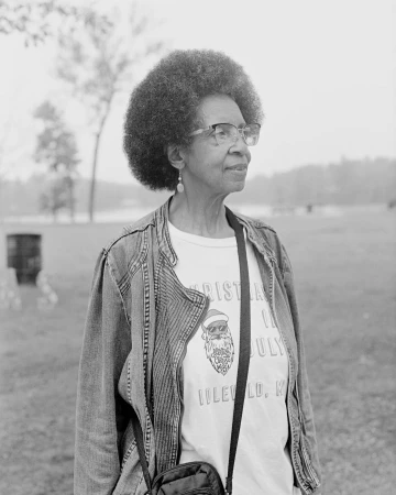Monochrome portrait of an elderly Black woman wearing glasses, a patterned t-shirt, and a jacket outdoors in New York.