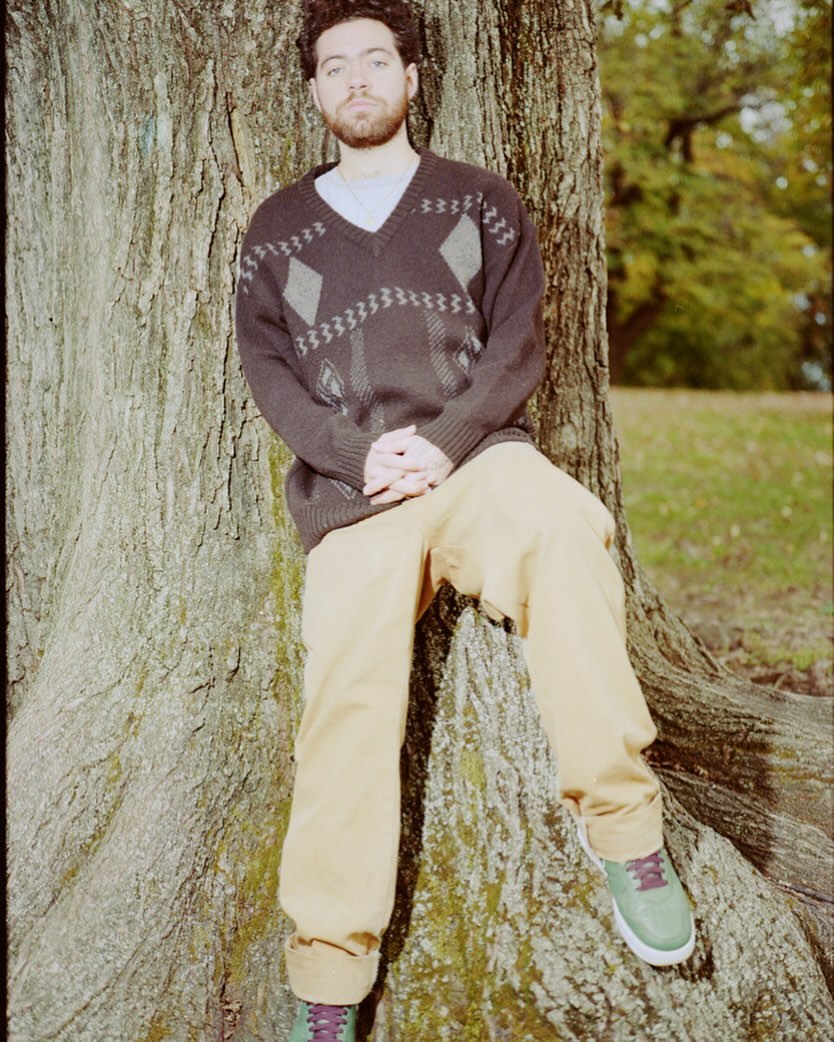 Adult male with a beard leans against a large tree wearing a patterned sweater and casual pants in an outdoor wooded environment.