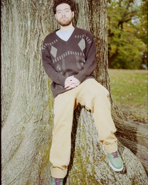 Adult male with a beard leans against a large tree wearing a patterned sweater and casual pants in an outdoor wooded environment.