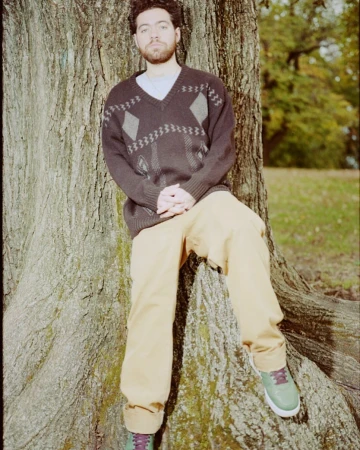 Adult male with a beard leans against a large tree wearing a patterned sweater and casual pants in an outdoor wooded environment.