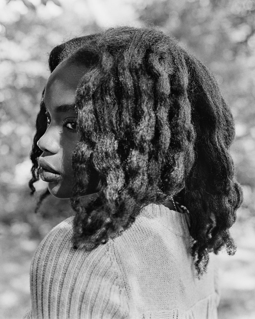 Black and white portrait of a young woman with curly hair looking over her shoulder in an outdoor setting with bokeh background.
