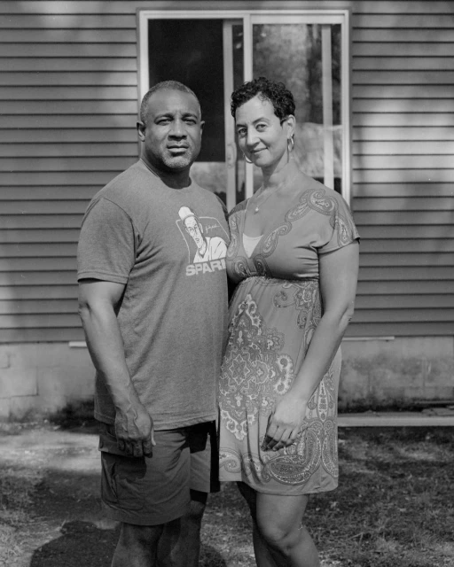 Portrait of an adult couple in casual summer clothing standing outside a house with a window on a sunny day in a black and white photograph.