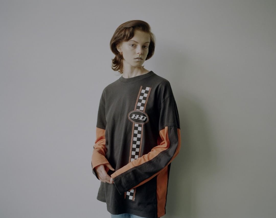 A young adult female poses indoors wearing a long sleeve graphic tee in a studio setting. The tee shirt has orange and black long sleeves.