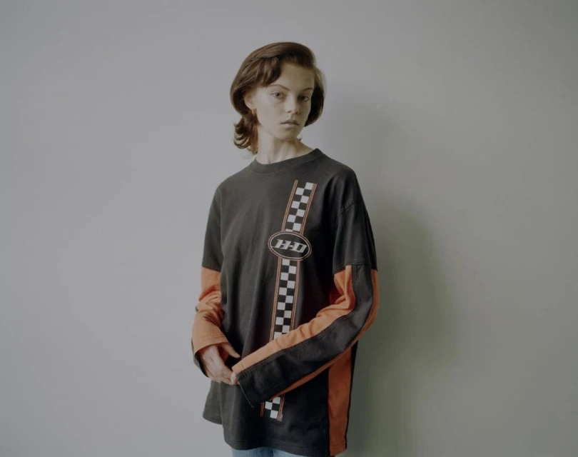 A young adult female poses indoors wearing a long sleeve graphic tee in a studio setting. The tee shirt has orange and black long sleeves.