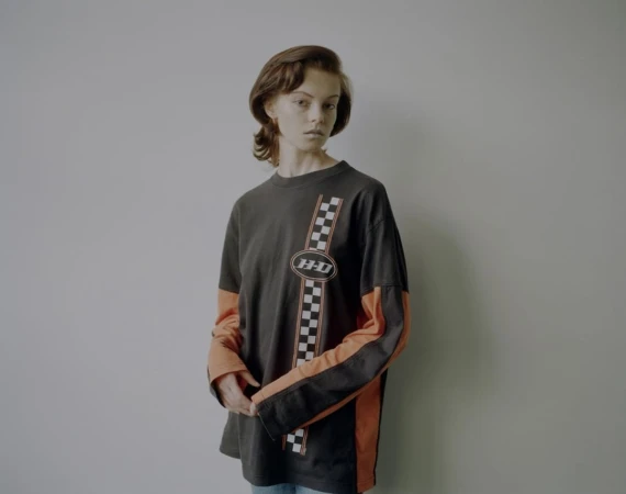 A young adult female poses indoors wearing a long sleeve graphic tee in a studio setting. The tee shirt has orange and black long sleeves.