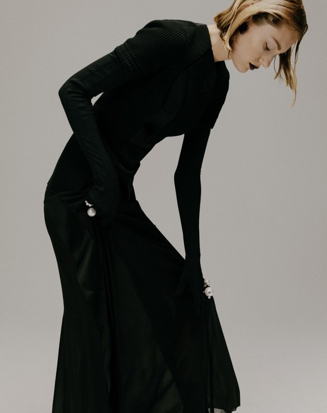 A slender woman poses in a long-sleeved black dress with a side slit and pearl accents, set against a soft grey backdrop in a studio setting.