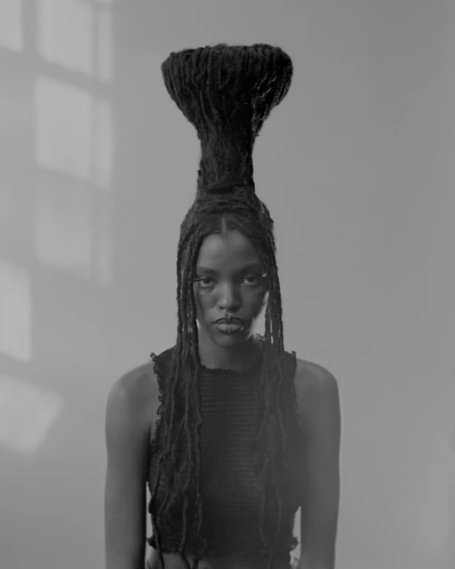 Monochromatic portrait of a Black woman with tall, sculpted dreadlocks, wearing a black top, set against a blurred grey background.