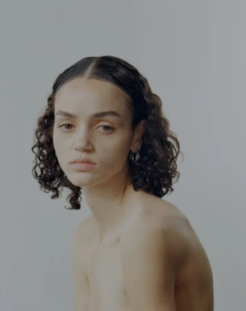 Portrait of a young woman with dark curly hair and hoop earrings against a neutral grey background by photographer Donavon Smallwood.