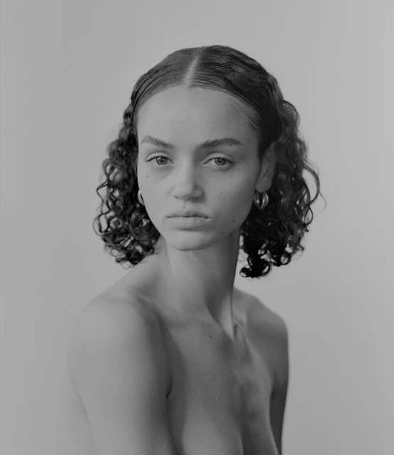 Monochrome portrait of a young woman with curly hair and hoop earrings, gazing directly at the camera against a light gray background.