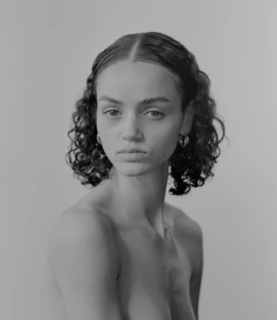 Monochrome portrait of a young woman with curly hair and hoop earrings, gazing directly at the camera against a light gray background.