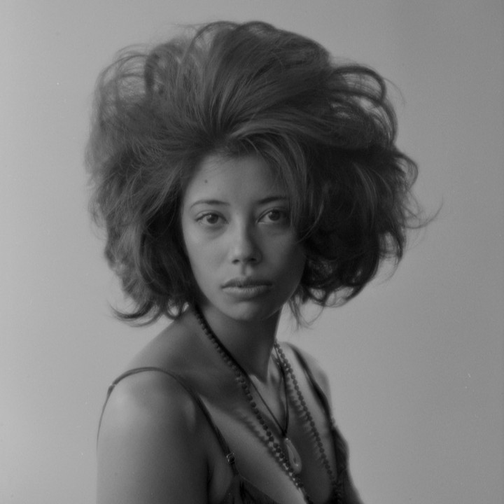 Monochrome portrait of a woman with voluminous hair, wearing a camisole and beaded necklaces, against a neutral grey background.