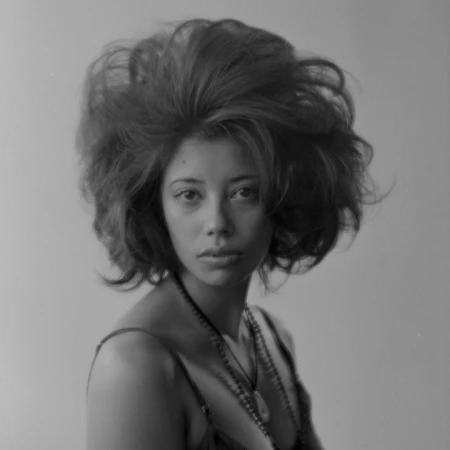 Monochrome portrait of a woman with voluminous hair, wearing a camisole and beaded necklaces, against a neutral grey background.