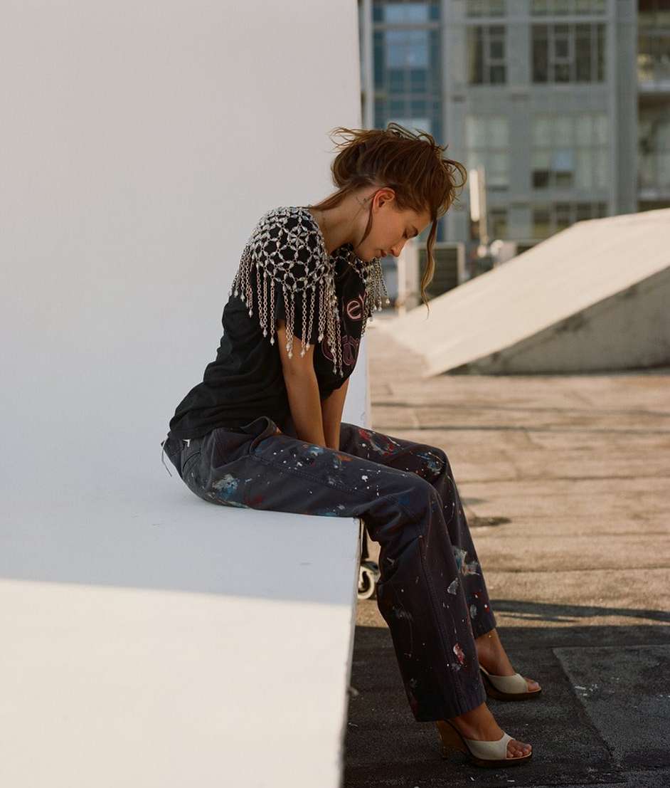 A woman with auburn hair sits on a rooftop wall in natural light, wearing a black t-shirt, paint-covered pants, and cream-colored heeled shoes.