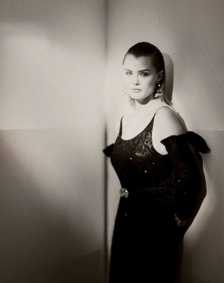 Elegant woman with shaved head in a black dress and earrings posing near a corner in a studio with soft, diffused lighting.