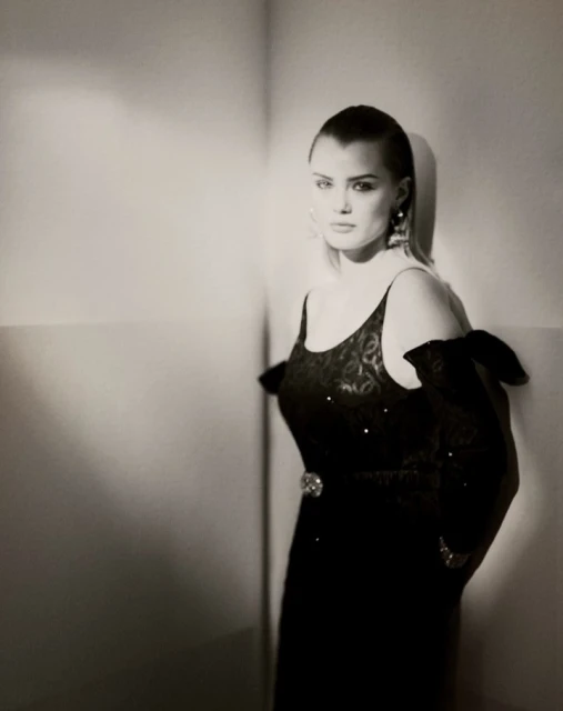Elegant woman with shaved head in a black dress and earrings posing near a corner in a studio with soft, diffused lighting.