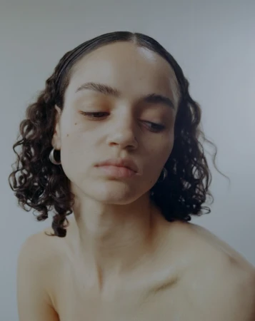 Portrait of a woman with dark curly hair and hoop earrings looking down against a neutral white background