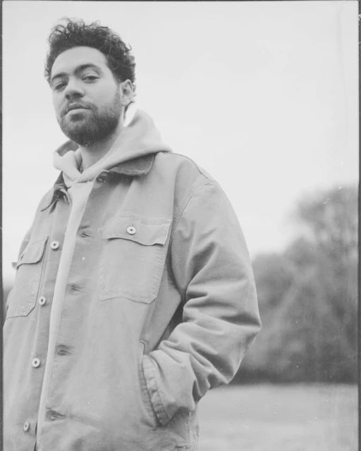 Portrait of a young adult male in outdoor setting, wearing a light brown jacket and light gray hoodie, posing with hand in pocket.