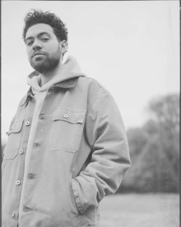 Portrait of a young adult male in outdoor setting, wearing a light brown jacket and light gray hoodie, posing with hand in pocket.