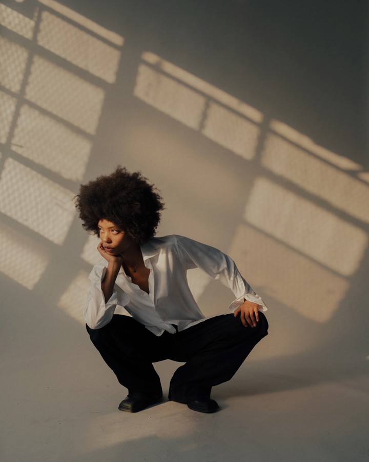 Young woman with afro in a white shirt and dark pants crouches indoors near a window with natural light casting shadows on the wall.
