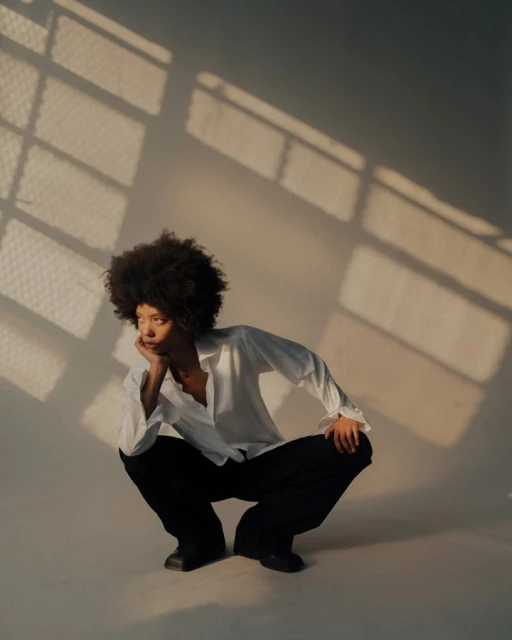 Young woman with afro in a white shirt and dark pants crouches indoors near a window with natural light casting shadows on the wall.