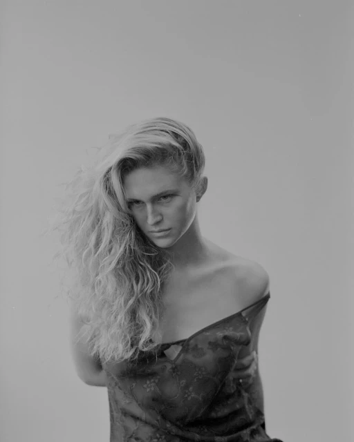 Monochrome portrait of a blond adult female model with curly hair and a dark floral pattern top, against a neutral studio backdrop.