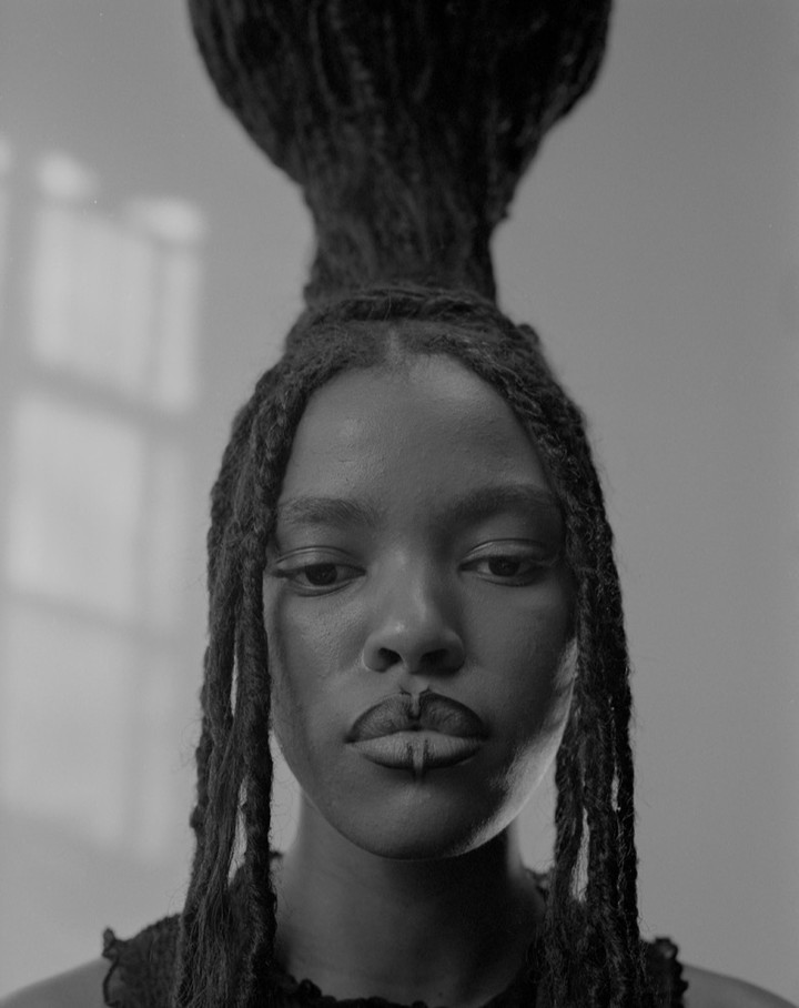 Monochromatic portrait of a Black woman with striking hairstyle and lip adornment, posing indoors with window light.