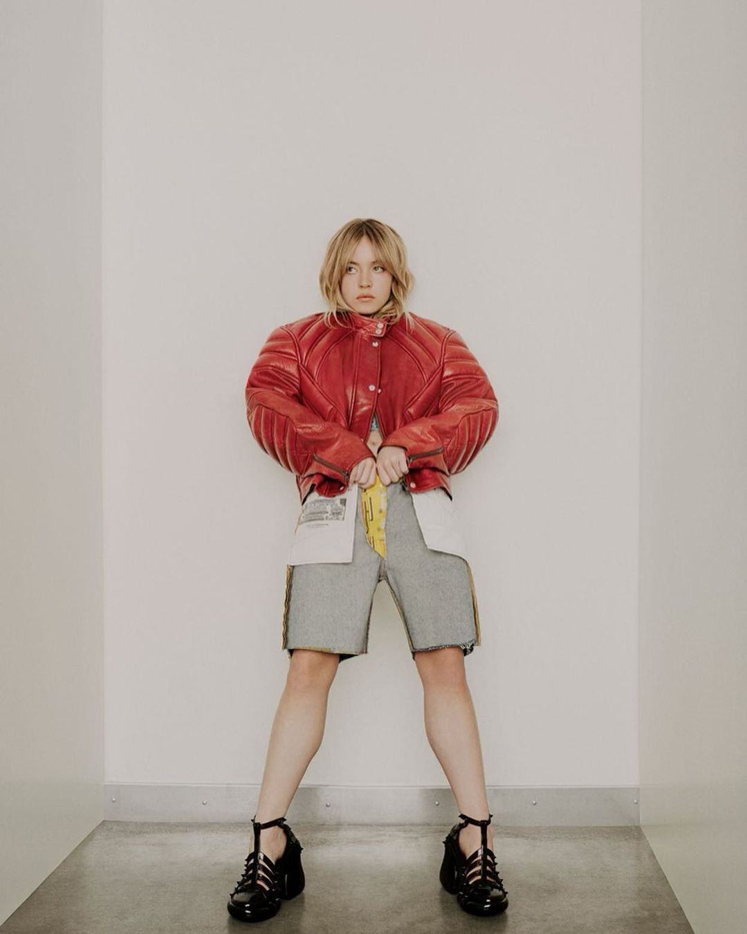 A full-length shot of a young adult woman in a red leather jacket, grey shorts, and black shoes standing in a bright white interior.