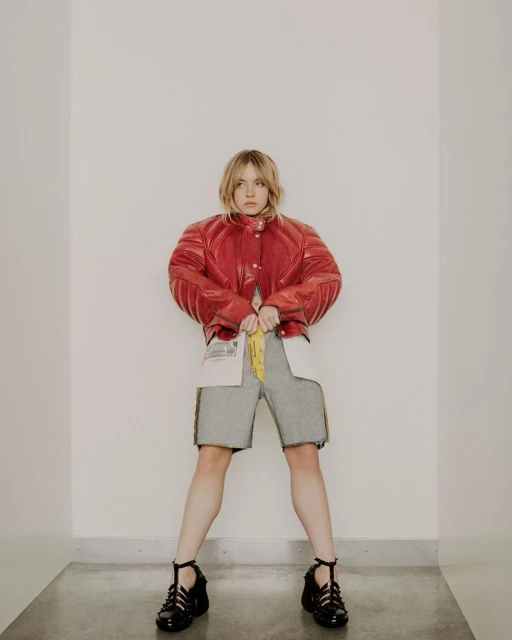 A full-length shot of a young adult woman in a red leather jacket, grey shorts, and black shoes standing in a bright white interior.