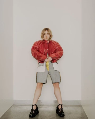 A full-length shot of a young adult woman in a red leather jacket, grey shorts, and black shoes standing in a bright white interior.