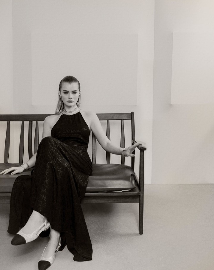 A model in a black halter dress and slingback heels sits on a wooden bench in a minimalist studio setting, showcasing fashion and style.