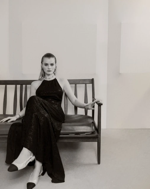 A model in a black halter dress and slingback heels sits on a wooden bench in a minimalist studio setting, showcasing fashion and style.
