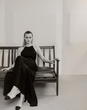 A model in a black halter dress and slingback heels sits on a wooden bench in a minimalist studio setting, showcasing fashion and style.