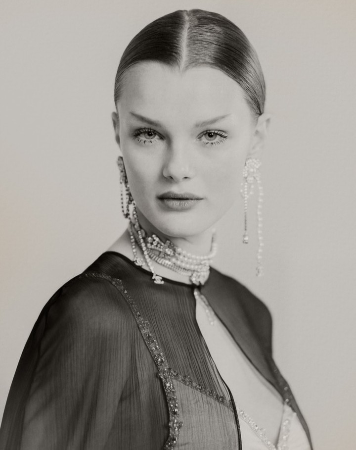 Close-up studio portrait of a woman with slicked-back hair, wearing a beaded choker, long earrings, and a sheer black capelet.