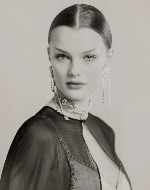 Close-up studio portrait of a woman with slicked-back hair, wearing a beaded choker, long earrings, and a sheer black capelet.