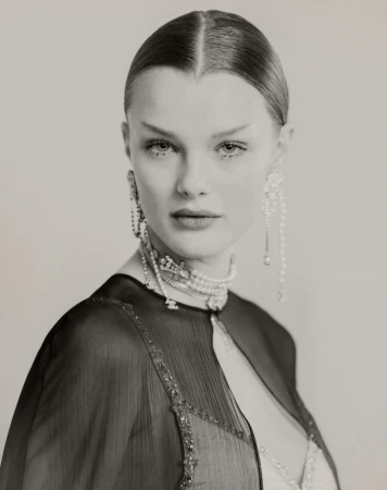 Close-up studio portrait of a woman with slicked-back hair, wearing a beaded choker, long earrings, and a sheer black capelet.