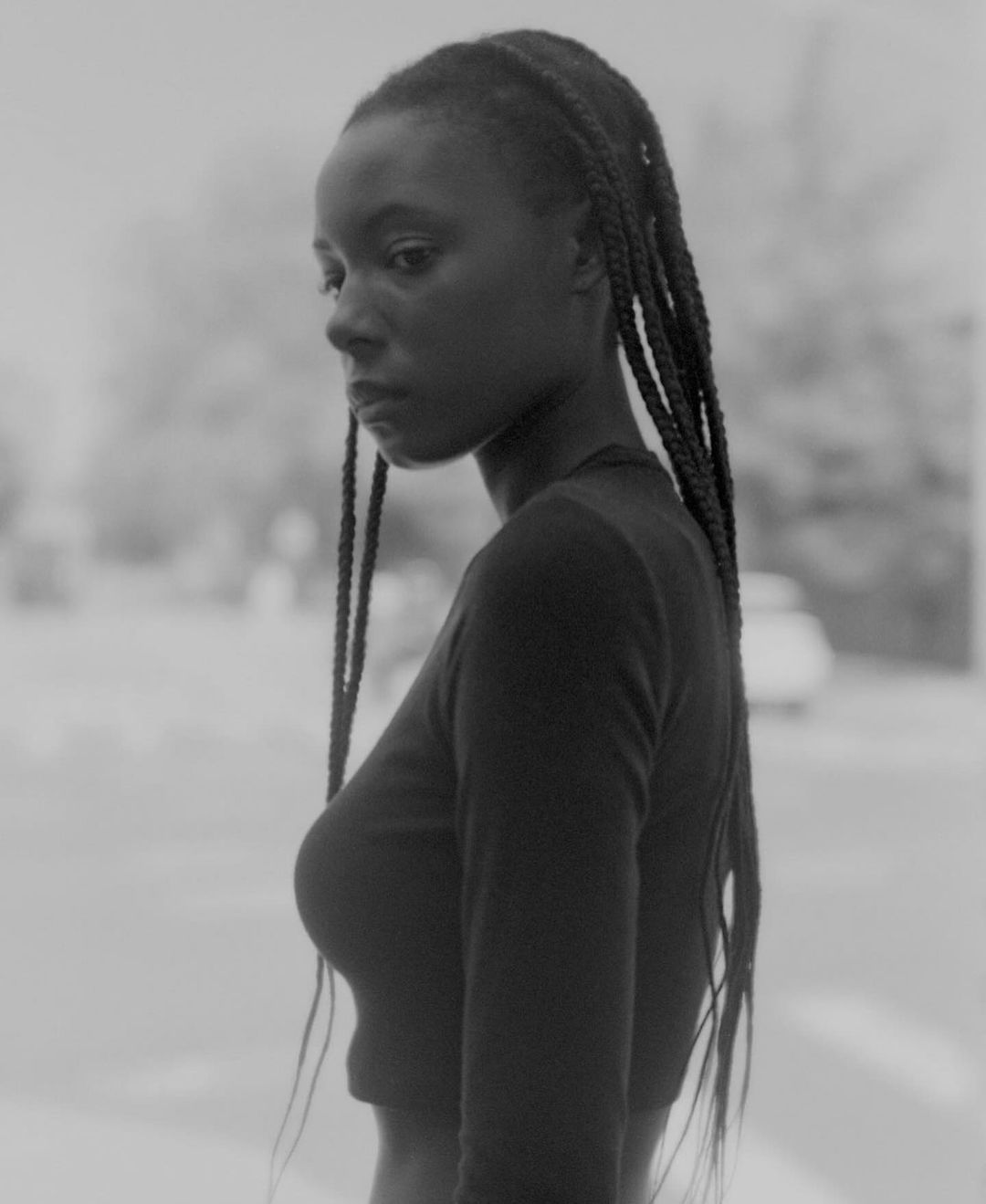 Monochrome portrait of a young adult African American woman with braided hair, wearing a black long-sleeve shirt in an urban setting.
