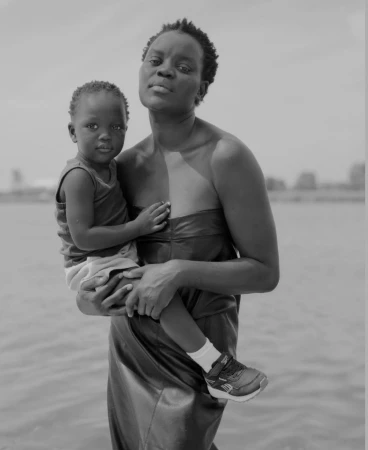Monochrome portrait of a black woman holding a young child outdoors near a body of water by Donavon Smallwood.