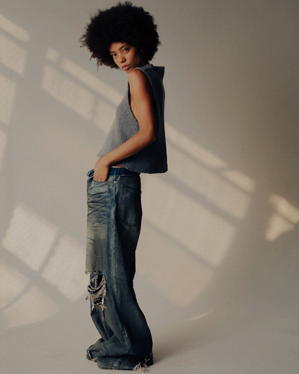 A young woman with afro-style hair poses in a photo studio, wearing a gray top and distressed, baggy jeans. Soft lighting creates shadows.